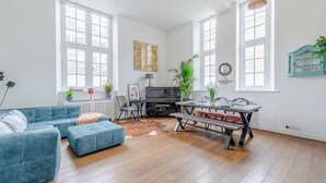 A spacious living room with a blue sofa, a dining table set for four, a piano, and large windows. - Luxurious & Chic 2BD Warehouse Flat - Old Street! (London)