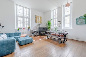 Un amplio salón con un sofá azul, una mesa de comedor preparada para cuatro personas, un piano y ventanas grandes.
