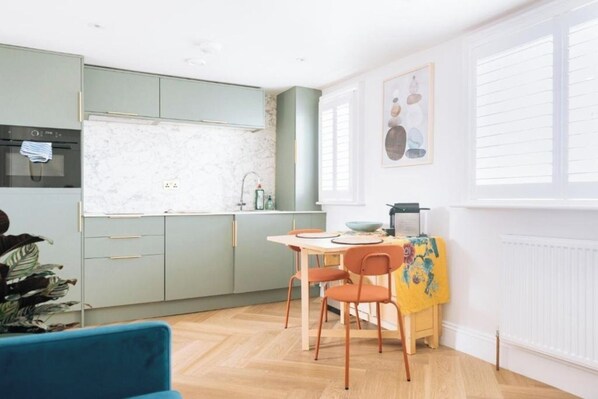 A modern kitchen with a dining area, featuring a herringbone floor pattern, a small dining table with chairs, and a window with blinds.