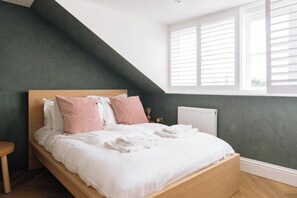 A bedroom with a sloped ceiling, a bed with white bedding and pink pillows, a wooden nightstand, and a window with white shutters. - Modern 1-Bed APT | Heart of Brighton & Sea Views (Brighton and Hove)