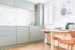 A modern kitchen with light grey cabinets, a marble countertop, a window with blinds, and a dining area with orange chairs.