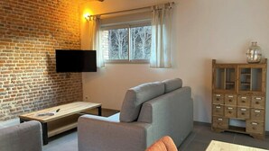 A cozy living room with a brick wall, a flat-screen TV, a sofa, a wooden cabinet, and a window with curtains.