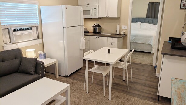 A compact kitchenette with a refrigerator, microwave, and stove. There is a small dining area with a table and chairs. The room has a neutral color scheme and a window with blinds.