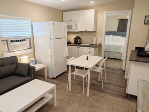 A compact kitchenette with a refrigerator, microwave, and stove. There is a small dining area with a table and chairs. The room has a neutral color scheme and a window with blinds.
