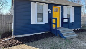 Una casa pequeña de un solo piso con fachada azul oscuro, contraventanas blancas y una puerta principal amarilla brillante.