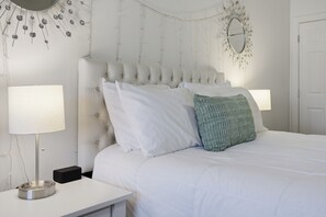 A neatly made bed with white bedding and a green throw pillow, flanked by two bedside tables with lamps, in a room with a tufted headboard and decorative wall hanging. - 5-Star Luxury Spacious 3BR/3BA Asheville Retreat • 5 Min to Downtown & Biltmore (Asheville)