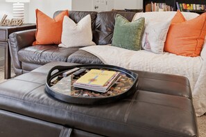 A living room with a leather sofa, a coffee table with a tray of magazines and a remote control, and a bookshelf in the background. - 5-Star Luxury Spacious 3BR/3BA Asheville Retreat • 5 Min to Downtown & Biltmore (Asheville)