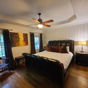 A bedroom with a large bed, a ceiling fan, a painting on the wall, and a window with curtains.