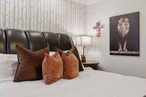 Een hotelkamer met een bed, bruine kussens, een hoofdbord, een lamp, een muur met een ingelijste afbeelding en een klein tafeltje met een decoratief object.