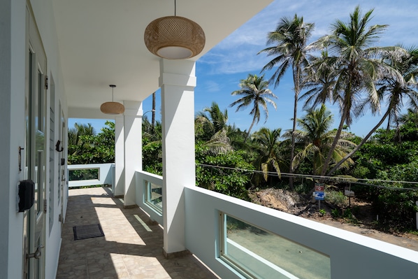 A balcony with white railings, a woven ceiling light, and a view of palm trees and greenery.