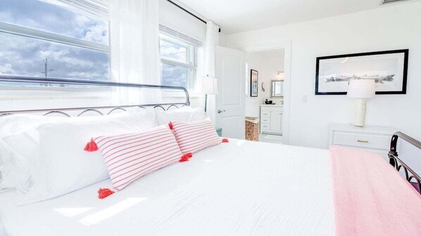 Un dormitorio con cama, ventana, puerta, mesita de noche y un cuadro enmarcado en la pared.