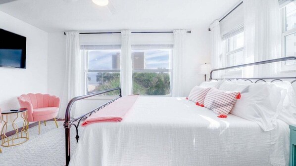 Una habitación de hotel con una cama, una silla rosa, una mesita y ventanas grandes.