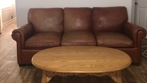 Ein braunes Ledersofa und ein Couchtisch aus Holz in einem Raum mit weißer Wand, einem gerahmten Bild und einem Regal mit Dekoration.