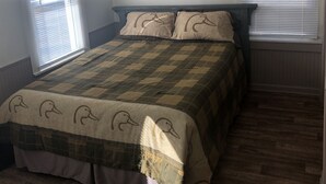 Ein Schlafzimmer mit kariertem Bettbezug, zwei Kissen mit Entenmotiven und einem Fenster mit Jalousien.