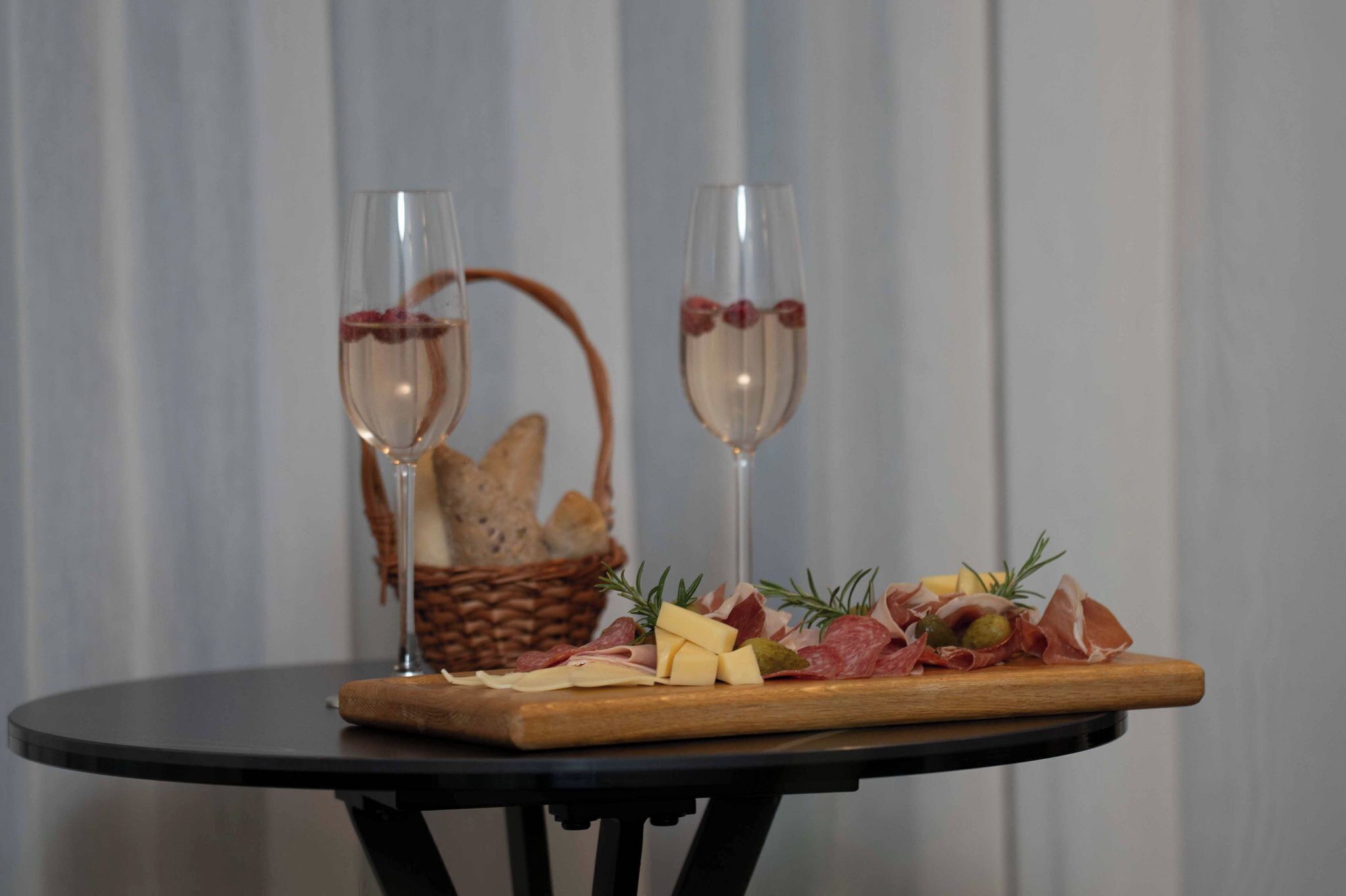A table with two glasses of wine, a wooden board with cheese and cured meats, and a basket of bread.
