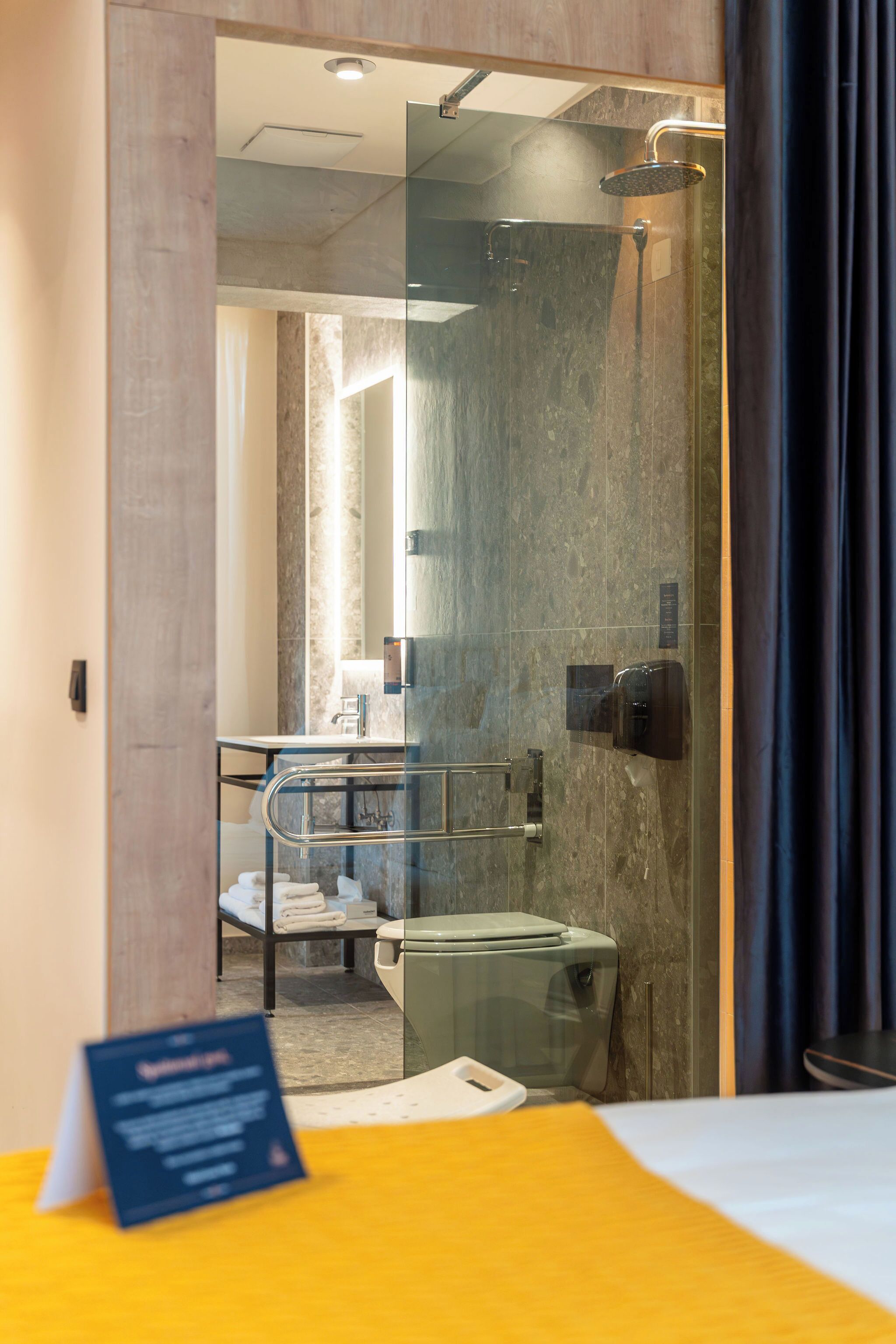 A modern hotel room with a glass shower enclosure, a bed with a yellow bedspread, and a visible bathroom with a toilet and towel rack.