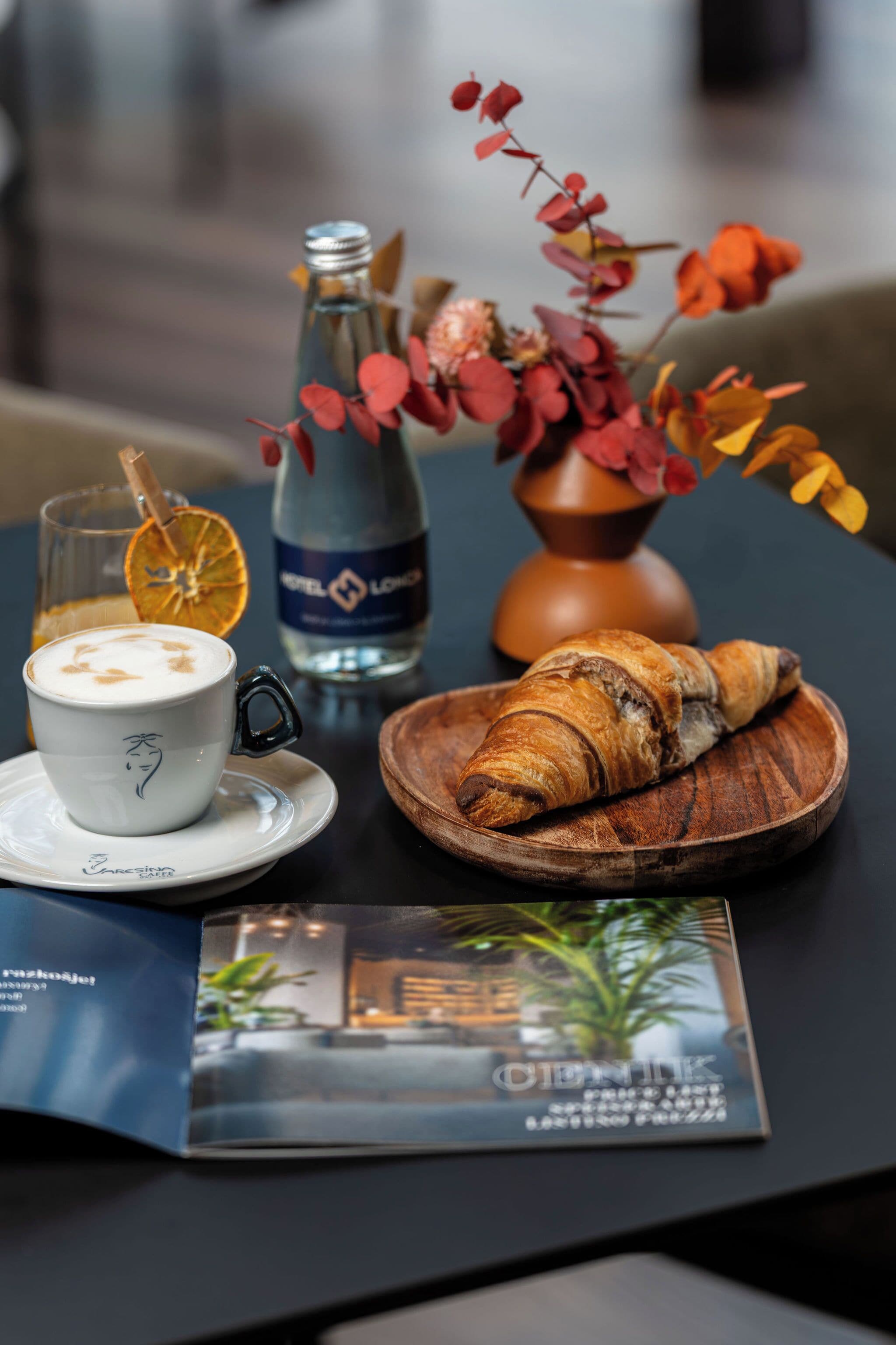 Una mesa con una taza de café, una botella de agua, un croissant, un plato de madera, un jarrón con flores secas y un folleto.