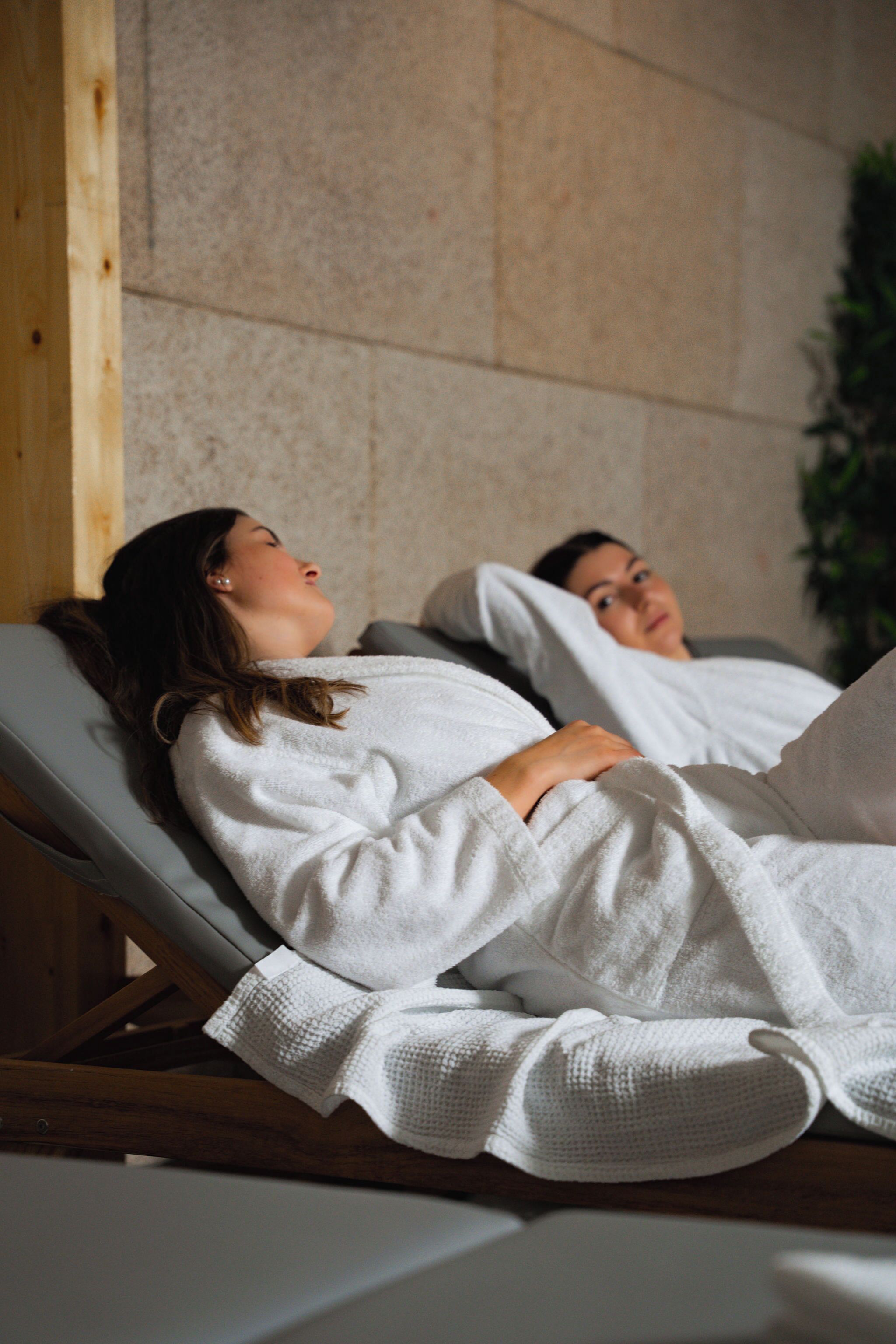 Two individuals lying on reclining chairs in a spa setting, covered with white towels.