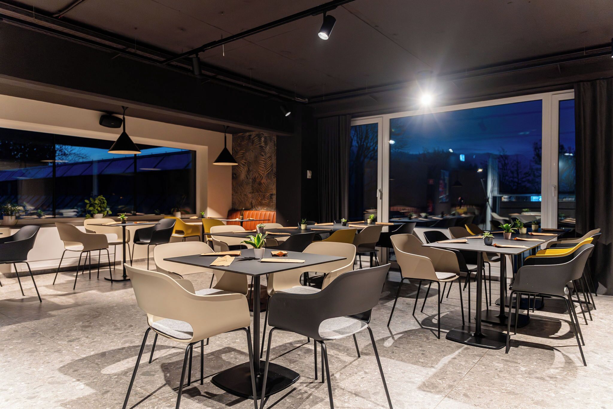 Un restaurante moderno con varias mesas y sillas, ventanas amplias y vistas a la ciudad al anochecer.