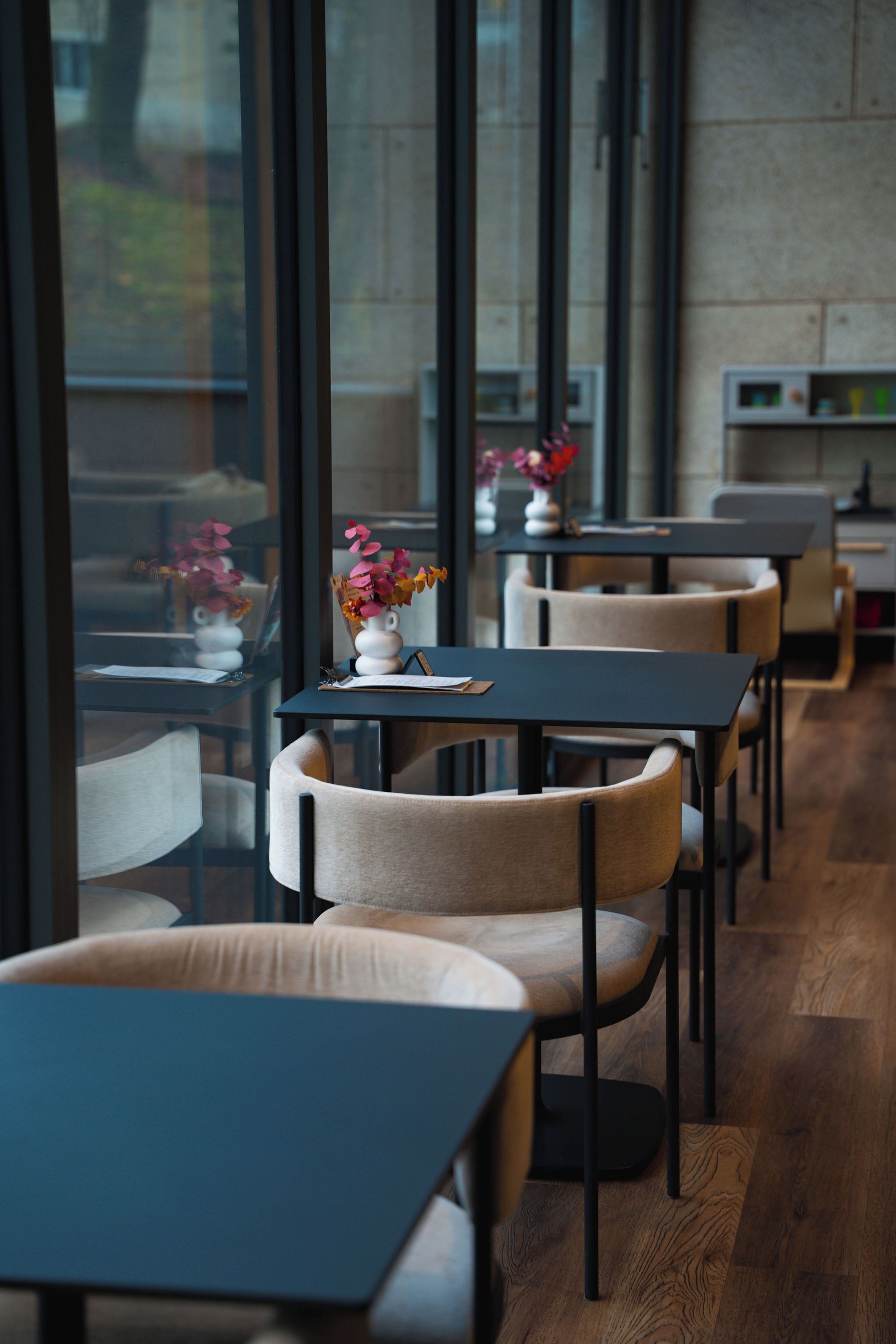 A modern restaurant interior with wooden flooring, black tables, and beige chairs.