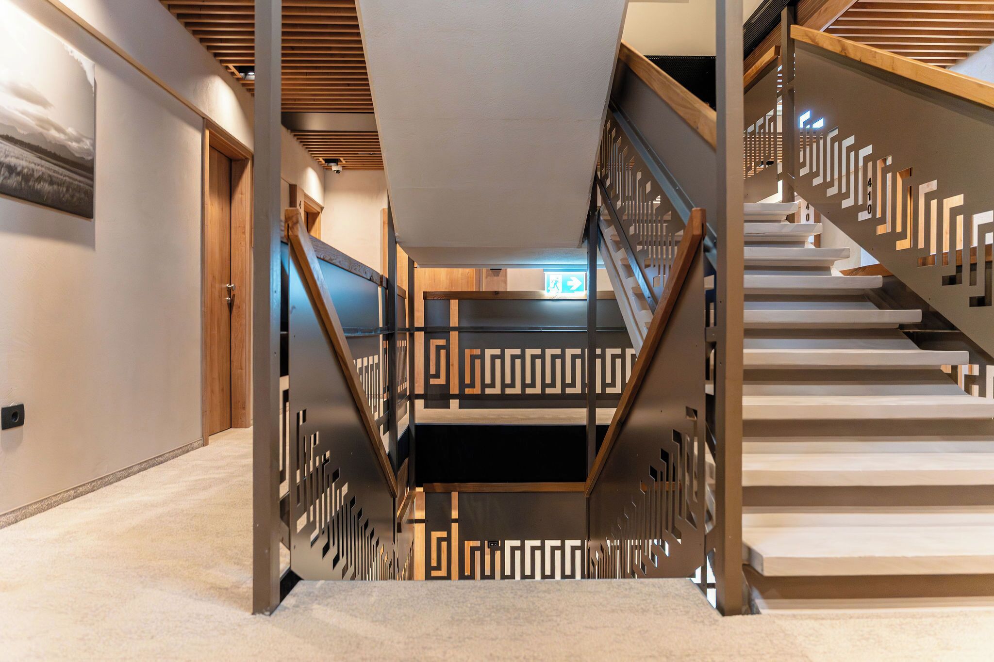 A modern interior with a wooden staircase, a ceiling with a geometric pattern, and a visible emergency exit sign.