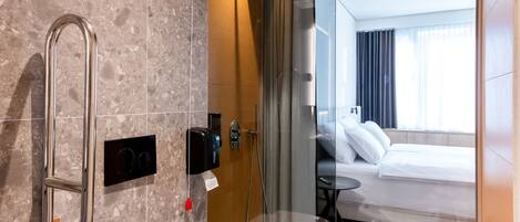 Un baño moderno con ducha, inodoro y un dormitorio con cama y ventana, separado por un cerramiento de vidrio.