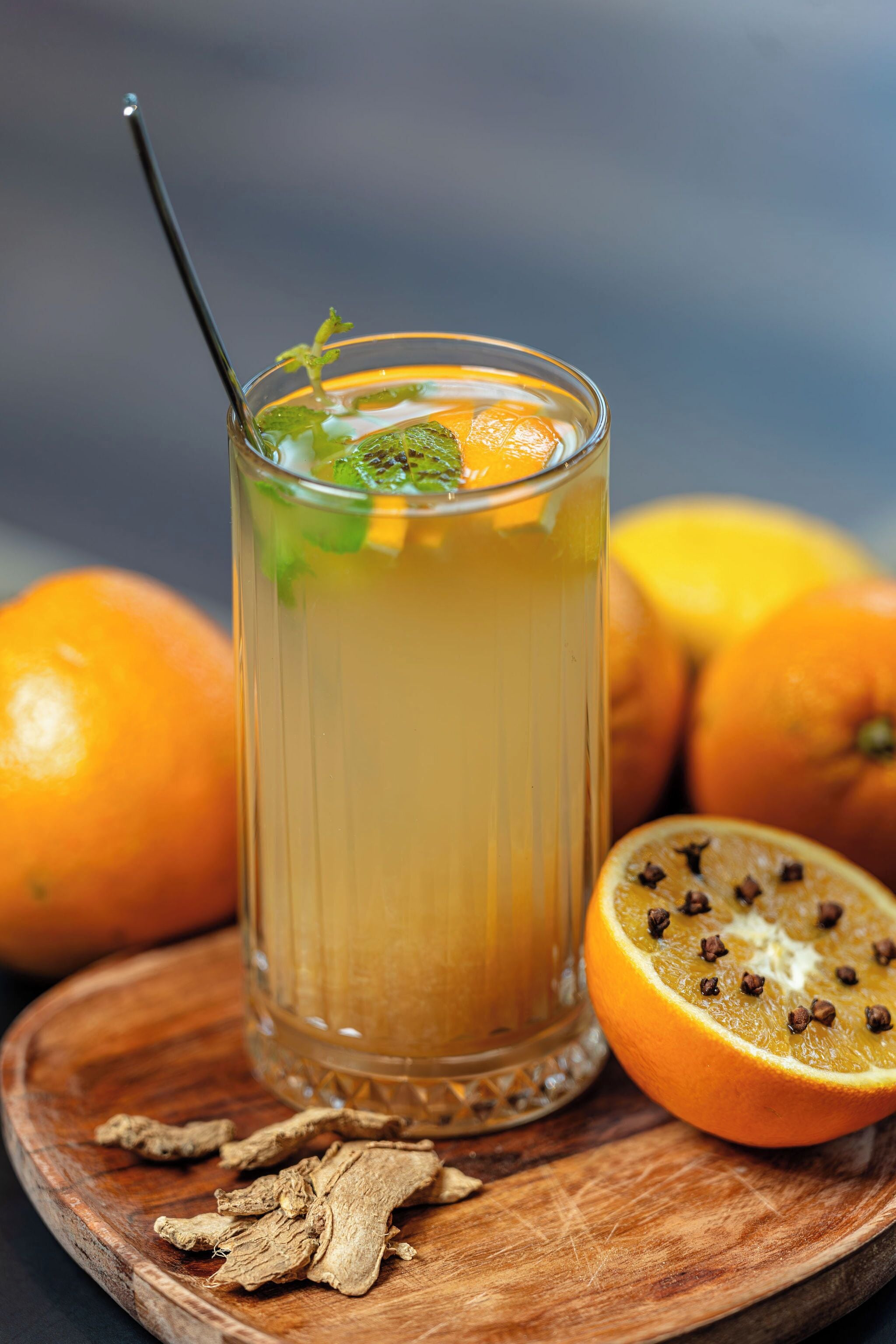 Un vaso de jugo de naranja con pajita, rodeado de naranjas y rodajas de jengibre sobre una tabla de madera.
