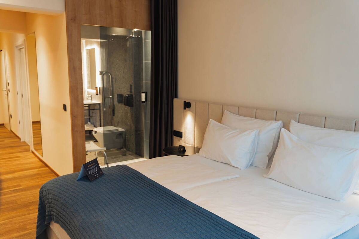 A hotel room with a bed, a nightstand, a bedside table, a glass shower enclosure, and a bathroom visible through the glass door.