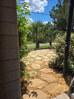 A stone pathway leading to a building entrance, surrounded by greenery and a palm tree.