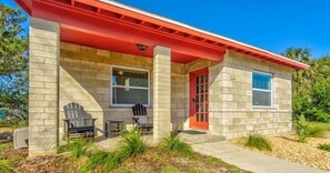 A small, single-story building with a red roof and beige walls. It features a front porch with two Adirondack chairs and a small table.