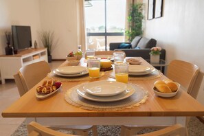 Une table à manger dressée avec des assiettes, des verres et des plats, dans une pièce bien éclairée où l’on aperçoit un téléviseur et des plantes en arrière-plan.