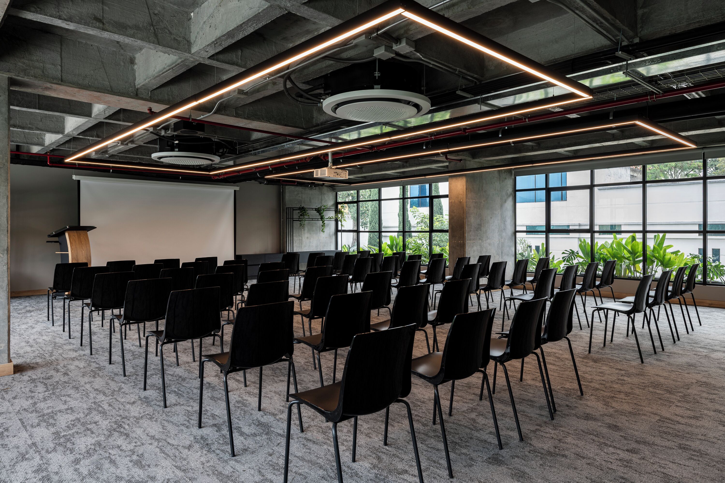A conference room with rows of chairs, a projector screen, and large windows with plants.
