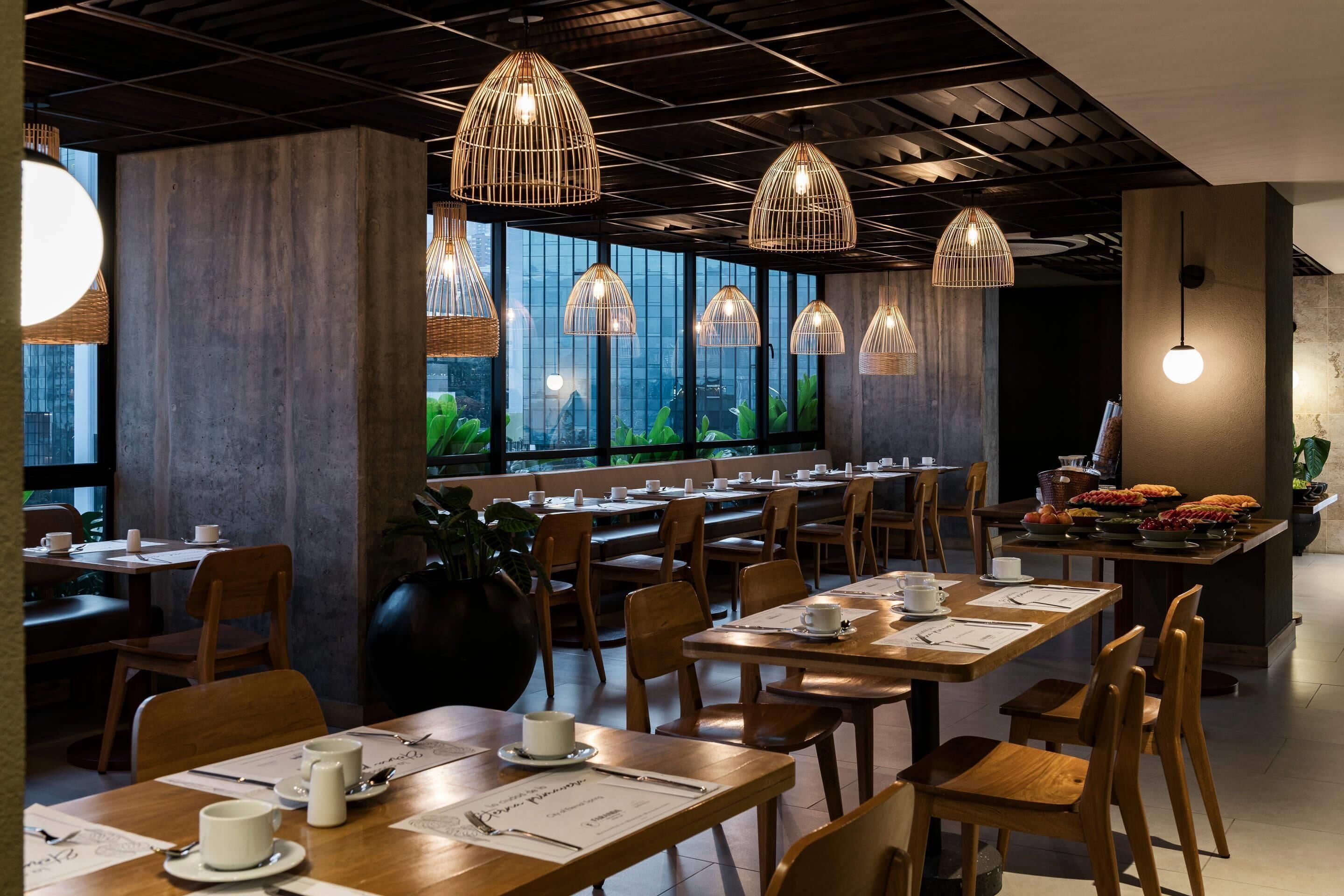 Un restaurante moderno con mesas y sillas de madera, lámparas colgantes y grandes ventanales que ofrecen vistas al exterior con vegetación.