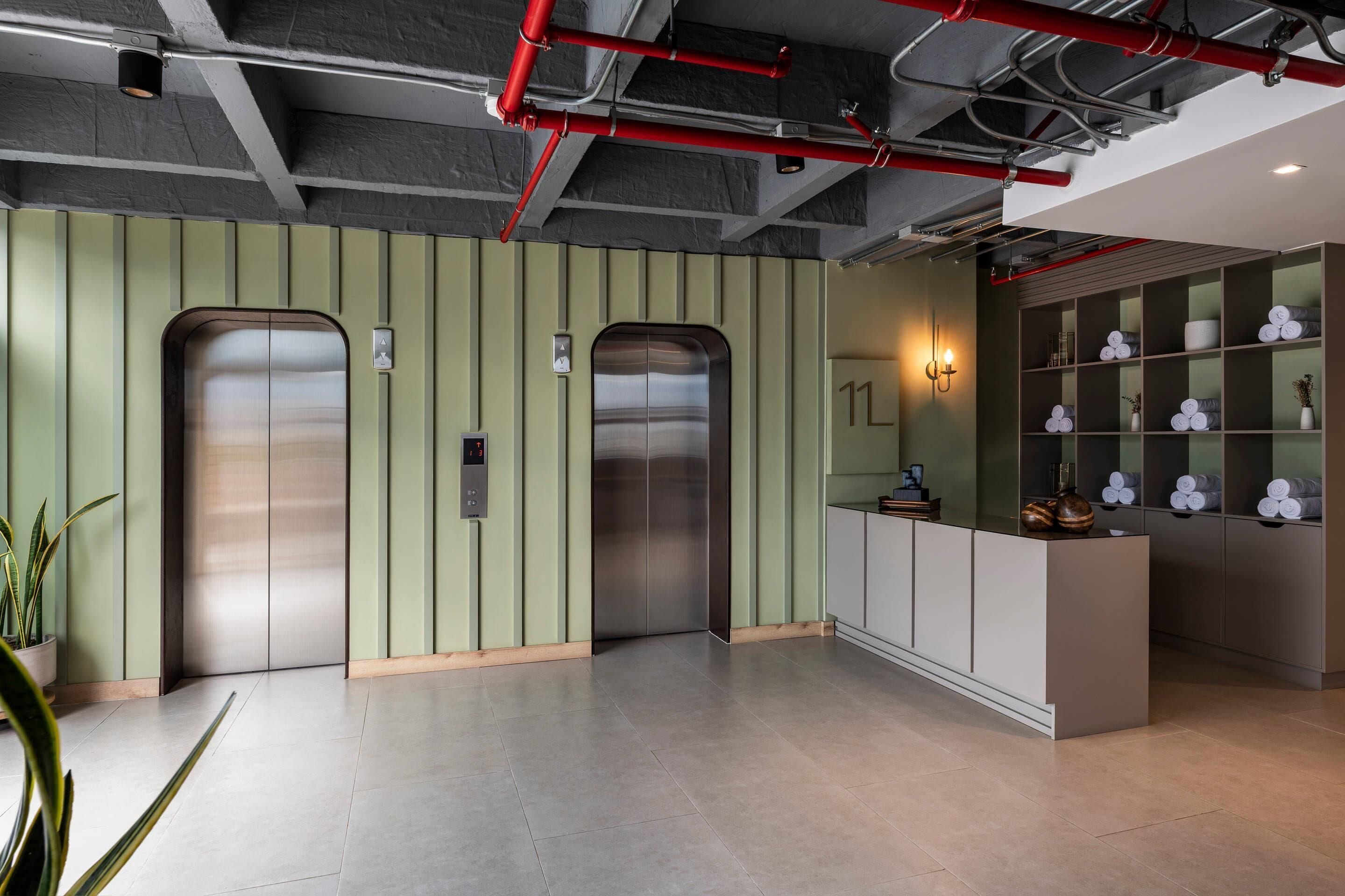 A modern lobby with two elevators, a reception desk, and a wall with shelves containing towels and toiletries.