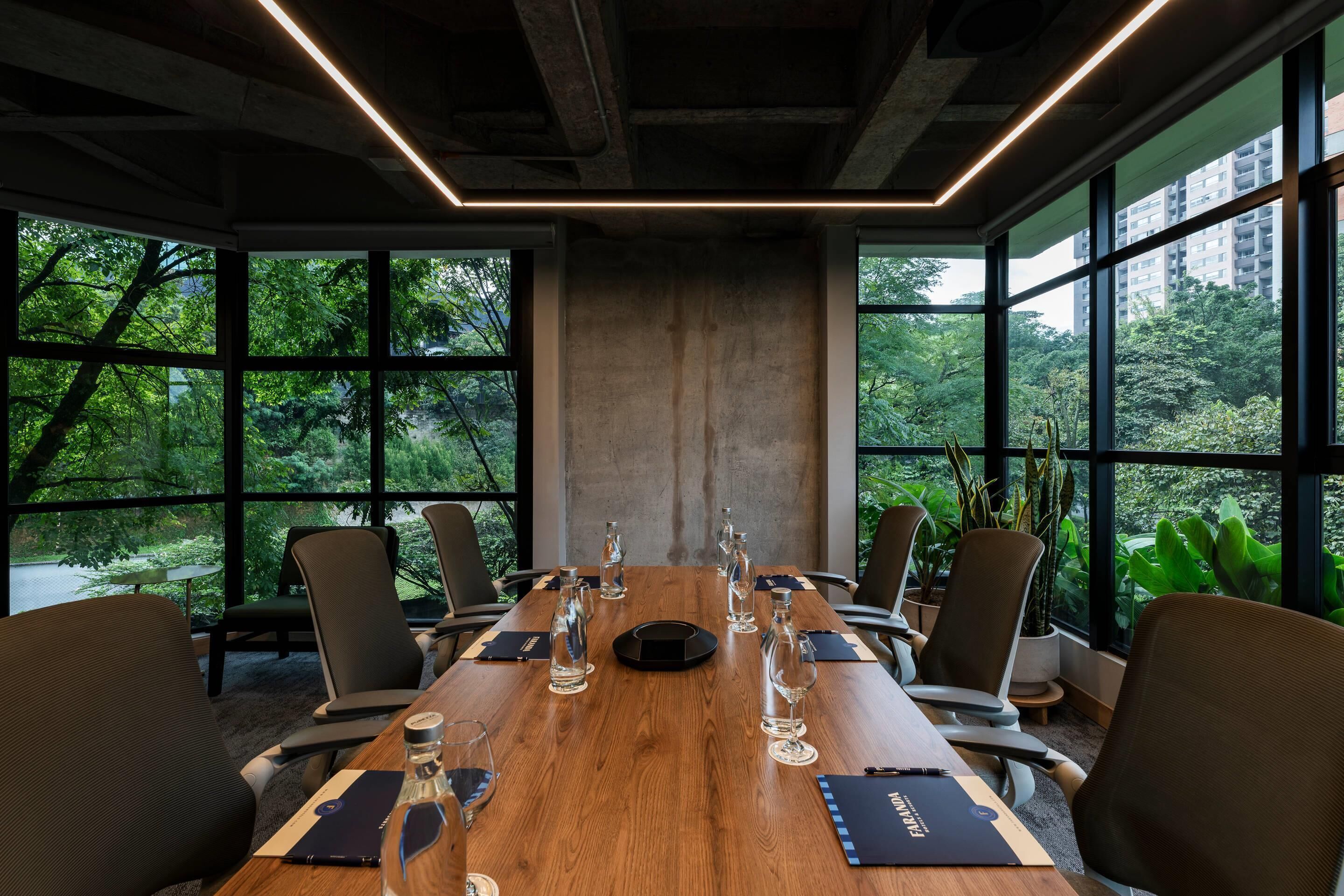 A conference room with a long wooden table, multiple chairs, and large windows offering a view of greenery.