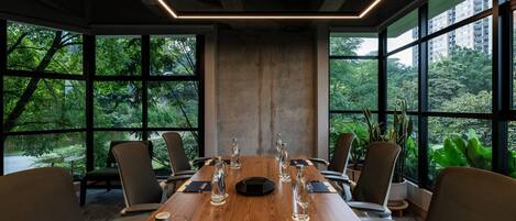 A conference room with a long wooden table, multiple chairs, and large windows offering a view of greenery.