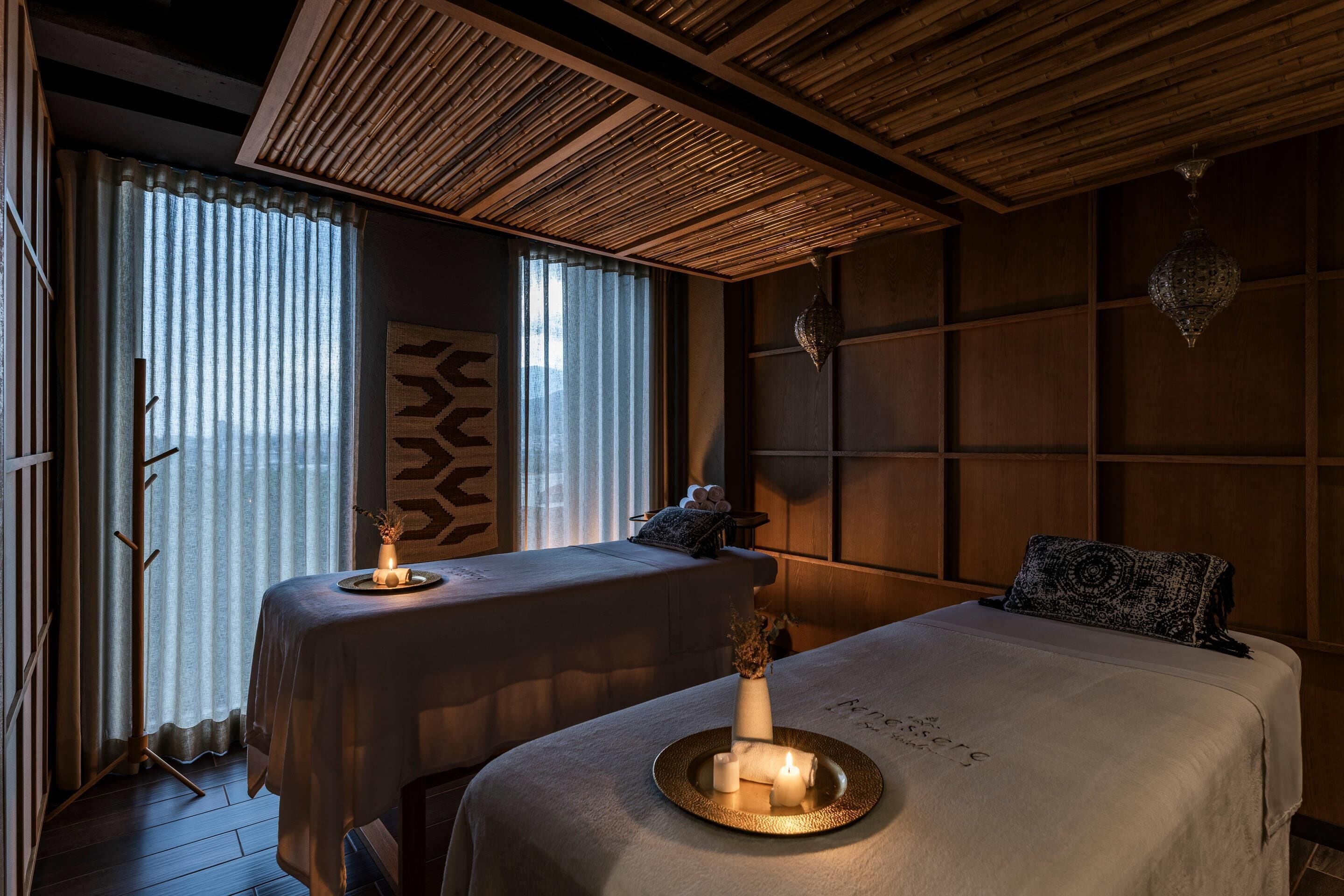 A room with two beds, a wooden ceiling, a large window with sheer curtains, and a decorative wall panel.