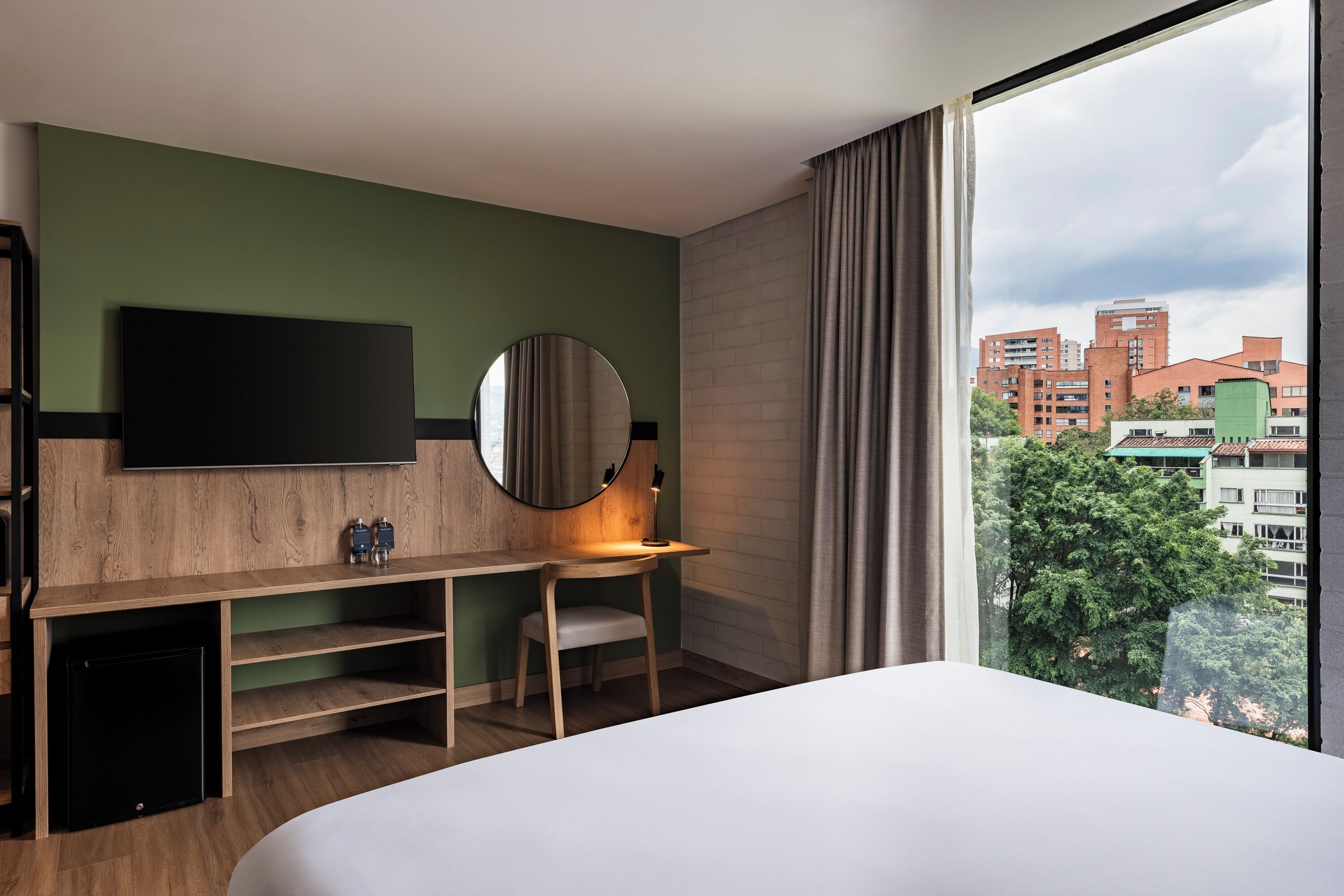 Habitación de hotel con cama, escritorio con silla, televisión y un ventanal con vistas a edificios y árboles.