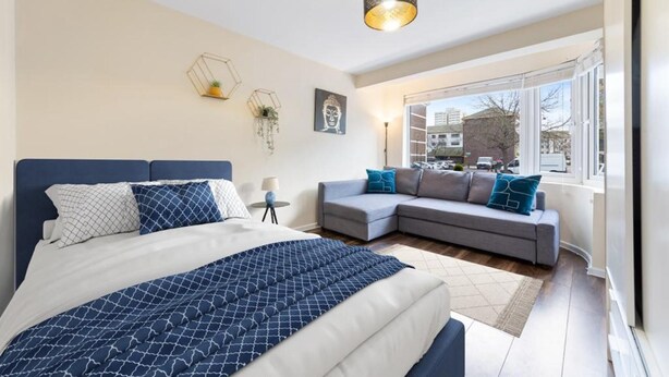 A modern bedroom with a bed, a grey sofa, and a window with a view of buildings. - Spacious Double Bed With Sofa Bed In Isleworth TW7 (Isleworth)