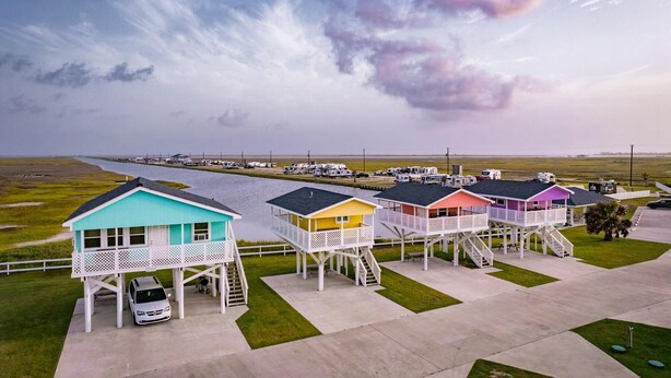 Una hilera de casas de playa coloridas con barandillas y escalones blancos, conectadas por un camino de hormigón, con vistas a la playa y un aparcamiento.