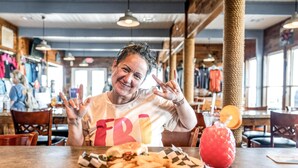 En person sitter vid ett bord på en restaurang och äter en hamburgare med pommes frites, och har ett glas framför sig. Restaurangen har träinredning och i bakgrunden syns klädställningar.