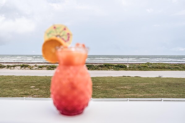 A drink in a glass with a slice of fruit on the rim, placed on a table with a view of the ocean and beach in the background.