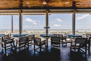 A covered outdoor seating area with wooden tables and chairs, a sign indicating a restroom, and a view of the beach and ocean.