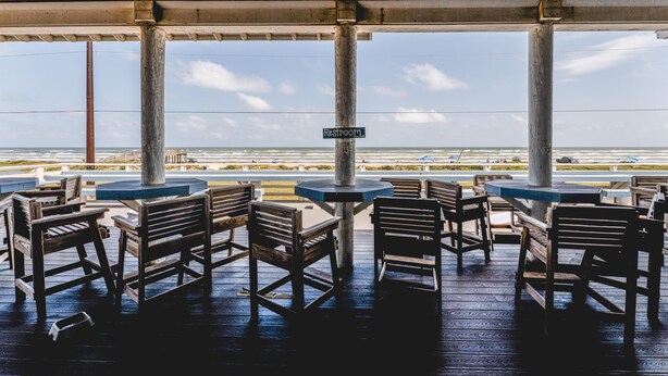 A covered outdoor seating area with wooden tables and chairs, a sign indicating a restroom, and a view of the beach and ocean. - The Pelican - Pelican - Two Bedroom Cottage (Freeport)