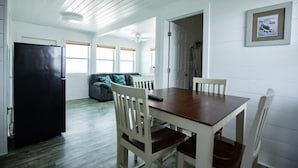 A dining area with a wooden table and chairs, a black refrigerator, a gray sofa with patterned cushions, and a framed picture on the wall. - The Pelican - Pelican - Two Bedroom Cottage (Freeport)