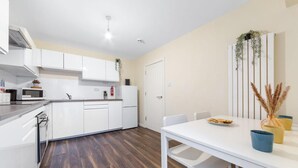 A modern kitchen with white cabinets, a microwave, and a refrigerator. There is a dining table with chairs and a vase with dried plants. - Stunning Double Bed Room In Isleworth TW7 (Isleworth)