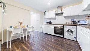 A modern kitchen with white cabinetry, a built-in oven, a microwave, and a washing machine. There is a dining area with a white table and chairs. - Stunning Double Bed Room In Isleworth TW7 (Isleworth)