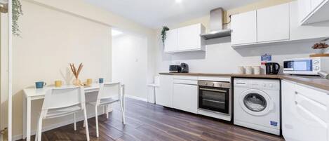 A modern kitchen with white cabinetry, a built-in oven, a microwave, and a washing machine. There is a dining area with a white table and chairs.