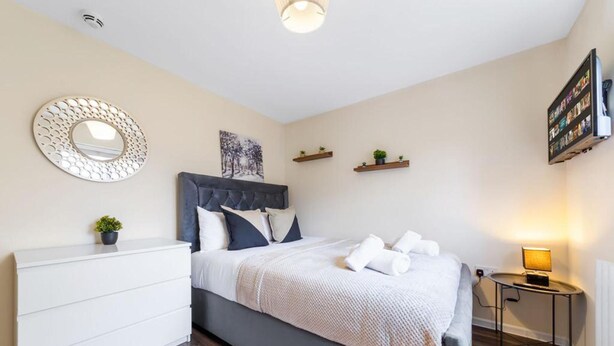 A bedroom with a bed, a dresser, a round mirror, a wall-mounted TV, and a small table with a lamp. - Stunning Double Bed Room In Isleworth TW7 (Isleworth)