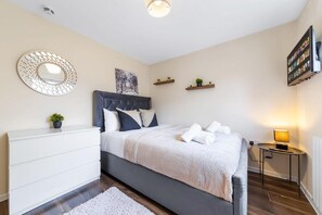 Une chambre à coucher comprenant un lit, une commode, un miroir rond, un téléviseur fixé au mur et une petite table avec une lampe.